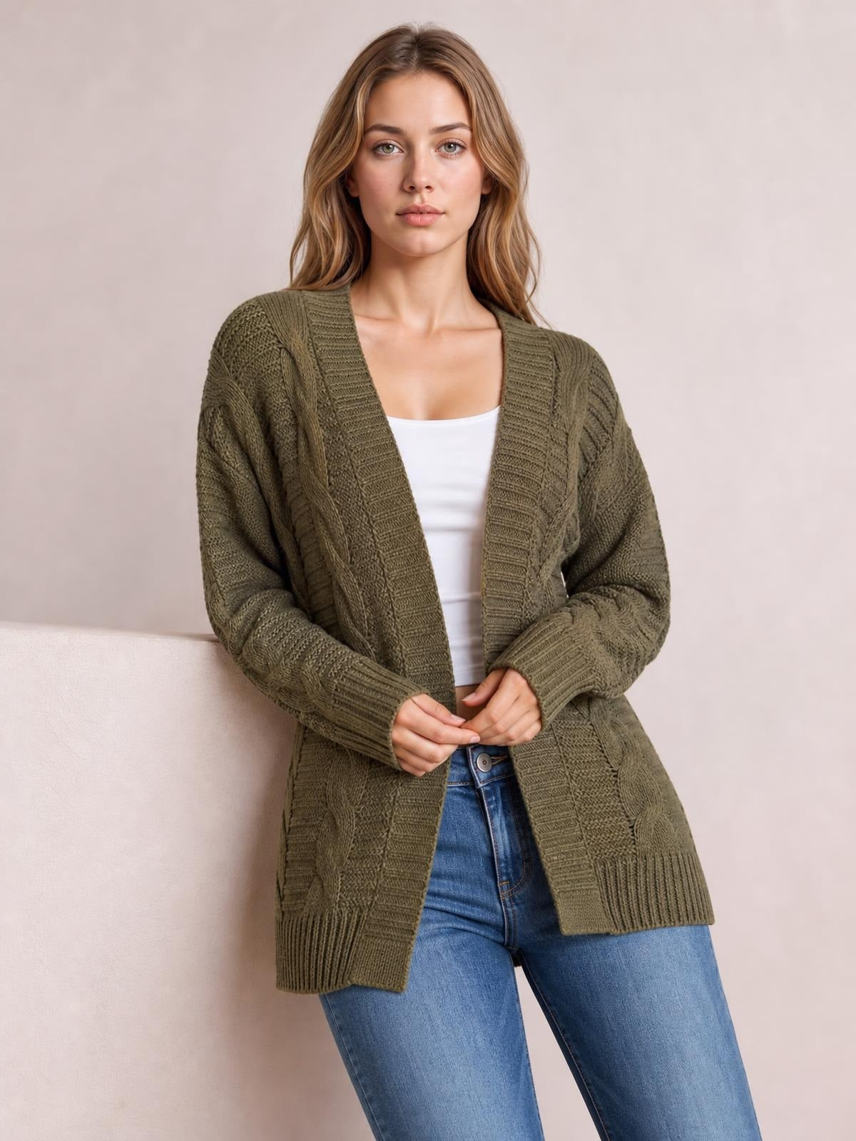 Model wears an olive green cable-knit open-front cardigan with long sleeves, ribbed trim, a relaxed longline silhouette, and hands clasped at the waist.