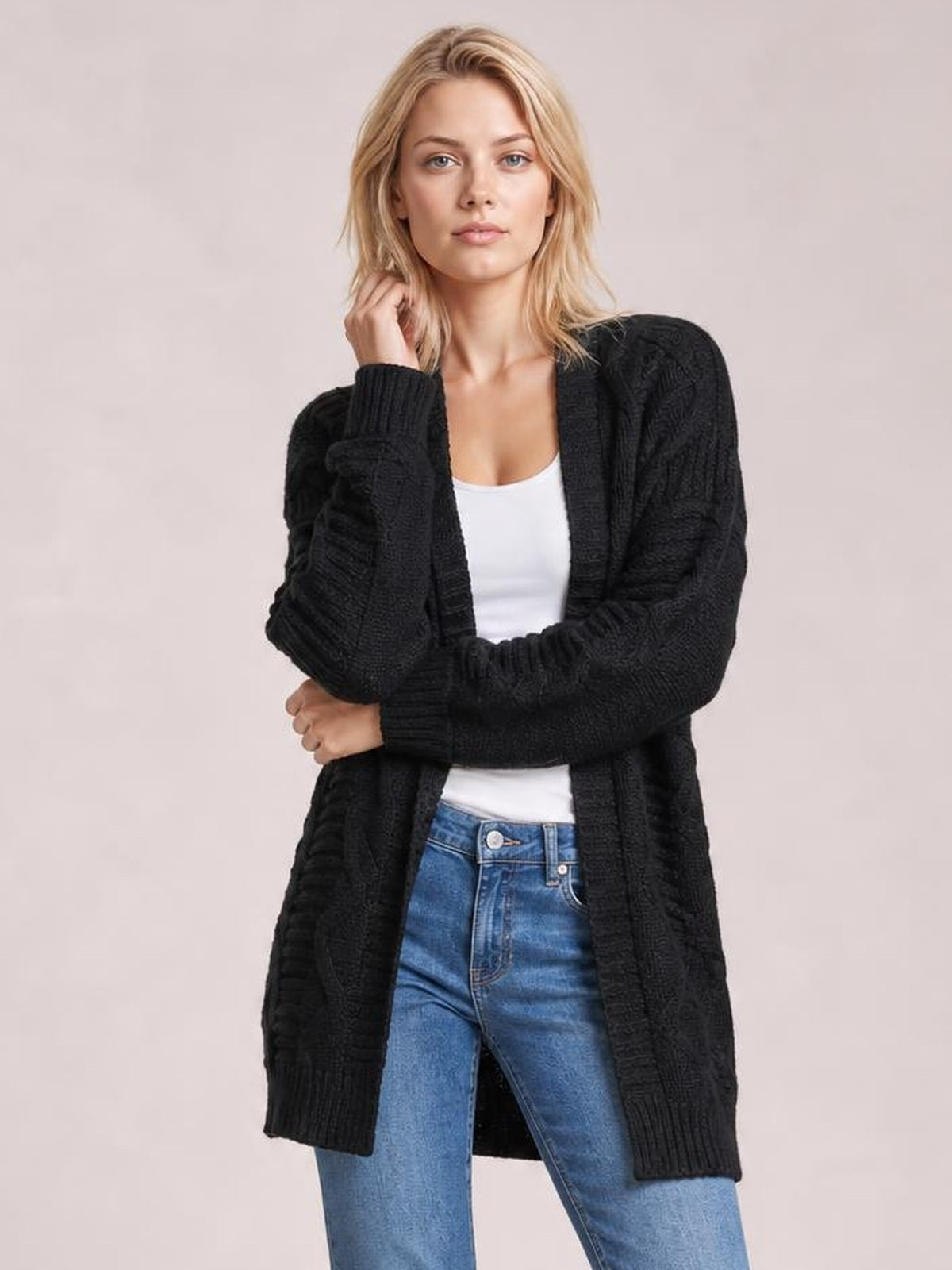 Model wears a black cable-knit open-front long-sleeve cardigan with a long relaxed silhouette and ribbed cuffs, posing with her arms crossed.