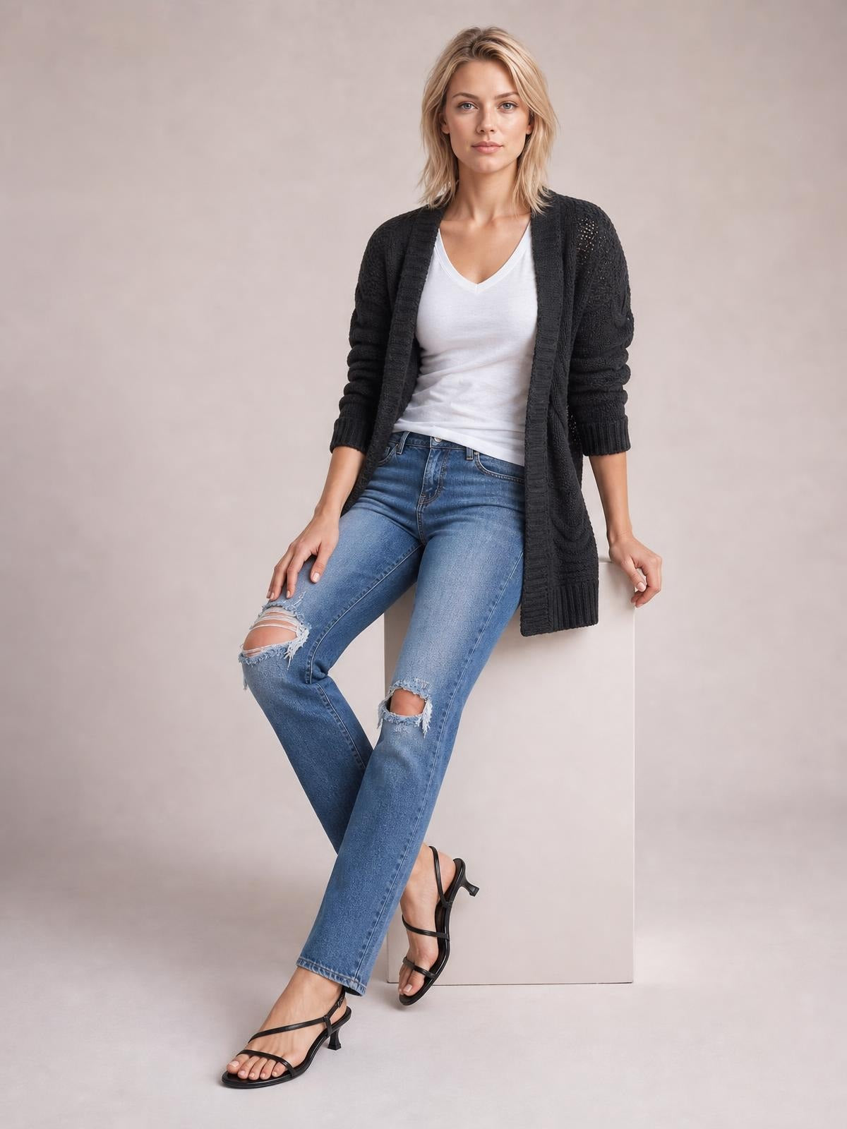 Model seated in a black cable-knit open-front long-sleeve cardigan with ribbed trim and a long relaxed silhouette over a white V-neck top.
