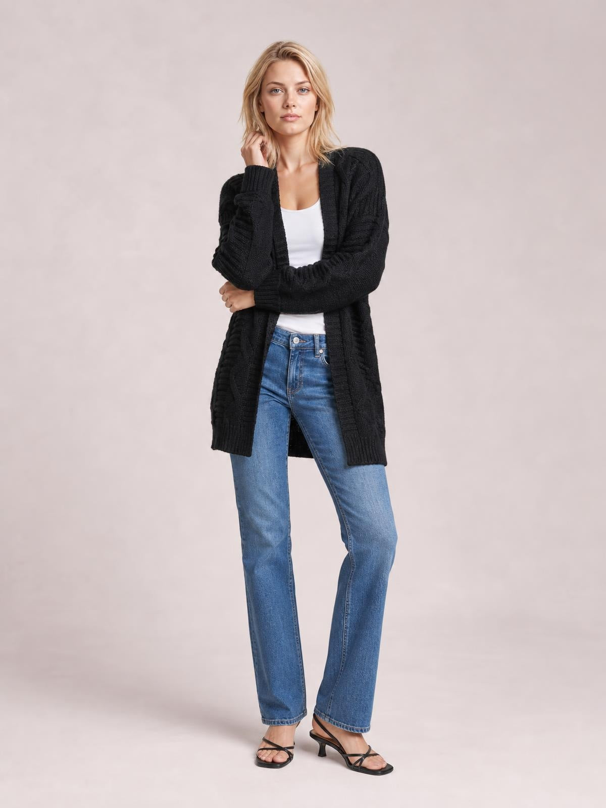 Model wears a black cable-knit open-front long-sleeve cardigan with a long relaxed silhouette, ribbed trim, and crossed arms over a white top.