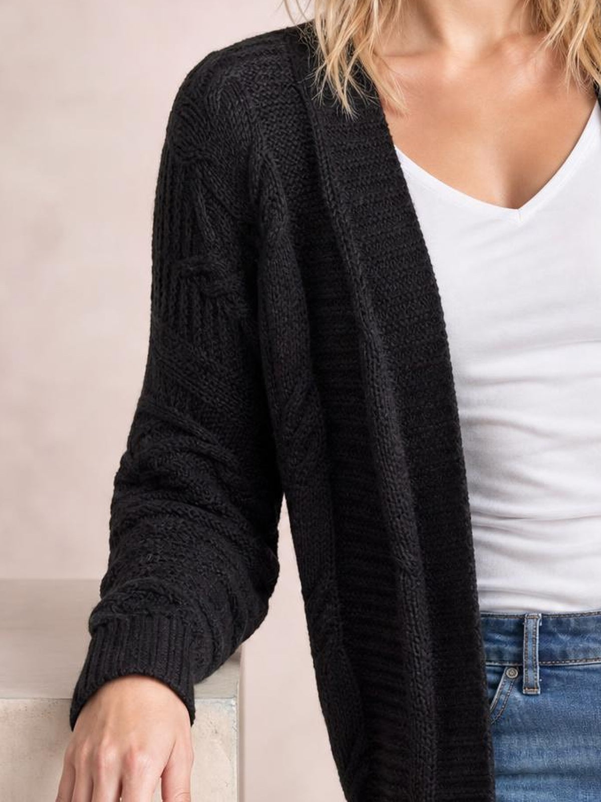 Model wears a black cable-knit open-front cardigan with long sleeves, ribbed cuffs, and a relaxed silhouette over a white V-neck top.