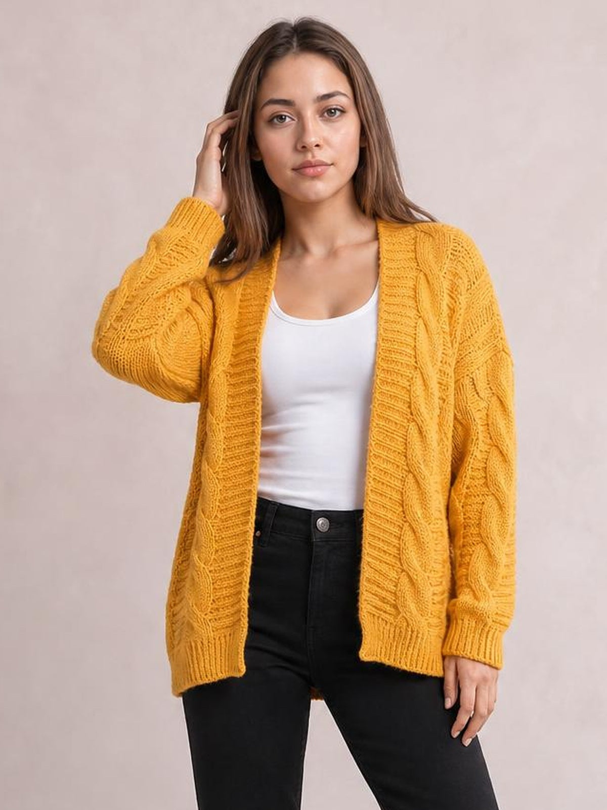 Model wearing a mustard cable-knit open-front cardigan with long sleeves, ribbed trim, an oversized relaxed silhouette, and one hand resting near her hair.