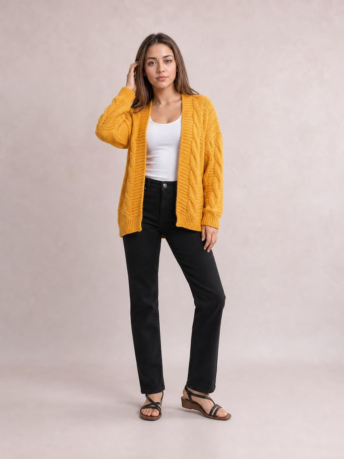 Model wearing a mustard-yellow cable-knit open-front long-sleeve cardigan with a relaxed silhouette and ribbed edges, posing with one hand in her hair.