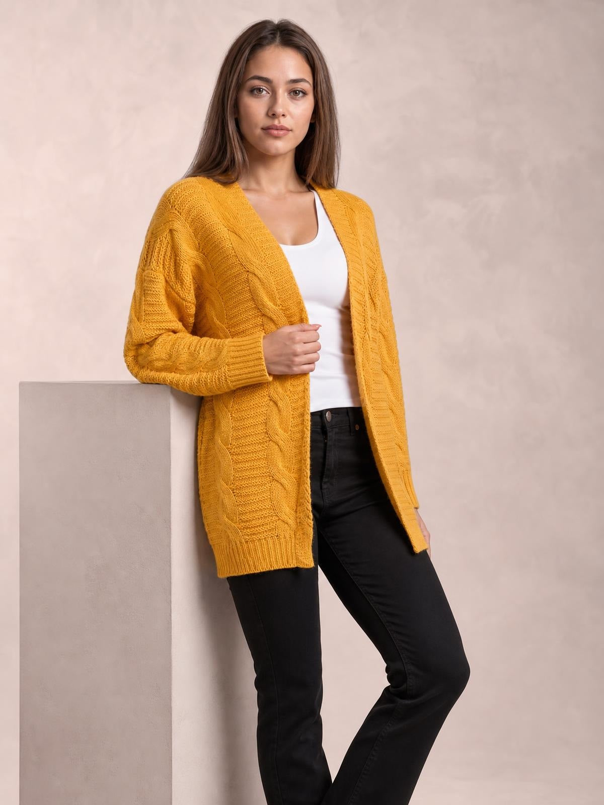 Model wearing a mustard yellow cable-knit open front long sleeve cardigan with a relaxed, longline silhouette over a white top and black pants.