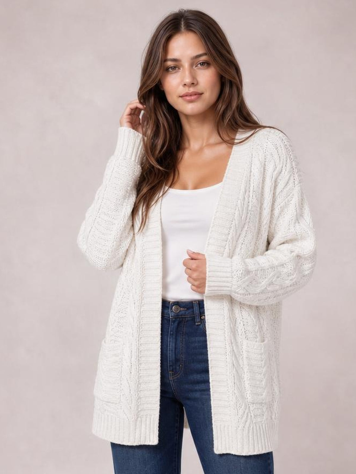 Model wears a white cable-knit open-front long-sleeve cardigan with a long relaxed silhouette, ribbed cuffs and hem, holding the front while touching her hair.