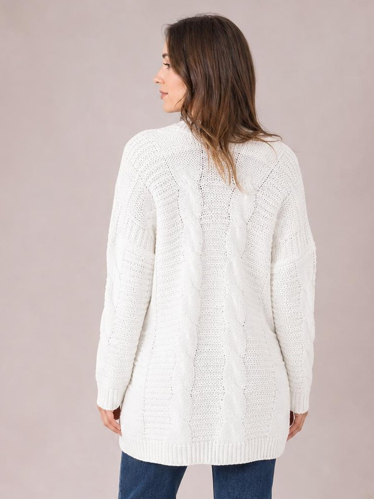 A model shown from the back wears a white cable-knit long-sleeve cardigan with a relaxed long silhouette, ribbed cuffs, and a ribbed hem.