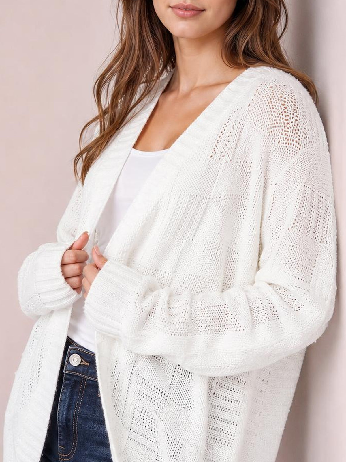 Model holds the front of a white cable-knit open-front cardigan, showing its relaxed silhouette, long sleeves, and textured knit construction over a white top.