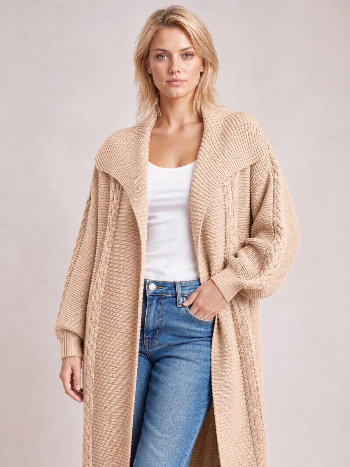Model wears a beige cable-knit open-front longline cardigan with ribbed trim, oversized sleeves, and a relaxed silhouette over a white top and jeans.