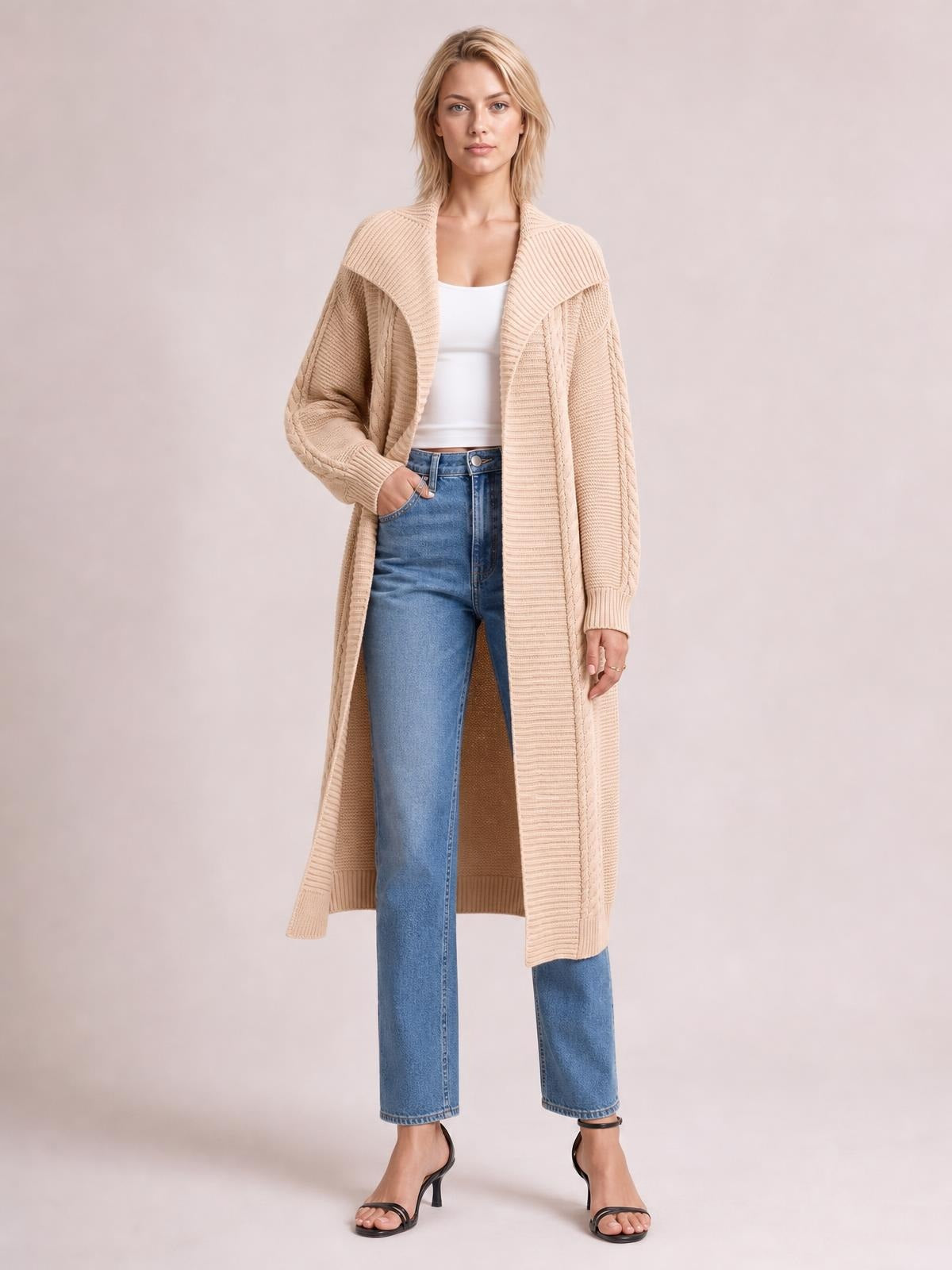 Model wears a beige cable-knit open-front longline cardigan with ribbed edges, draped in a relaxed silhouette over a white top and jeans.