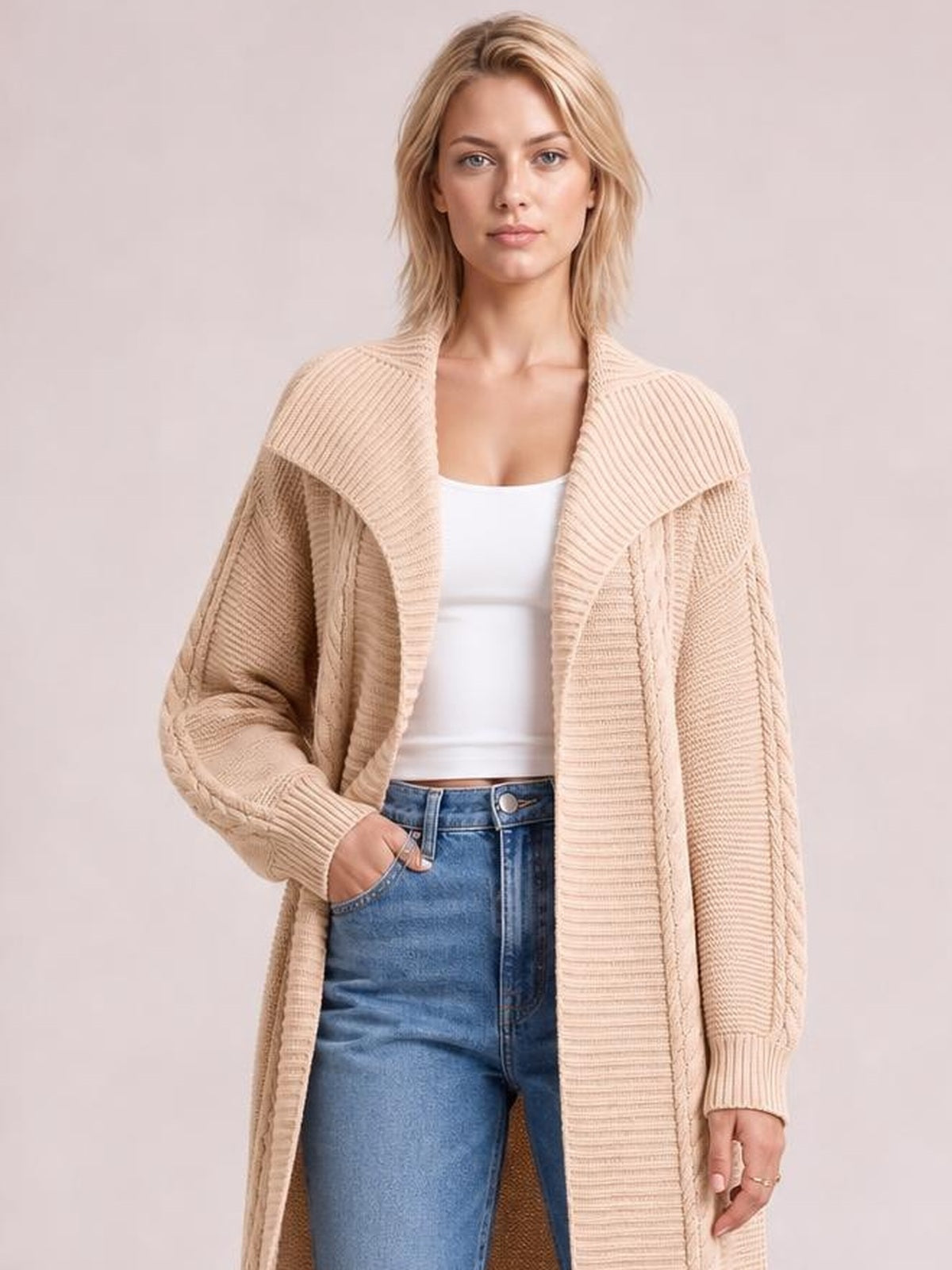 Model wearing a beige cable-knit open-front longline cardigan with ribbed trim, relaxed sleeves, and a long flowing silhouette over a white top and jeans.