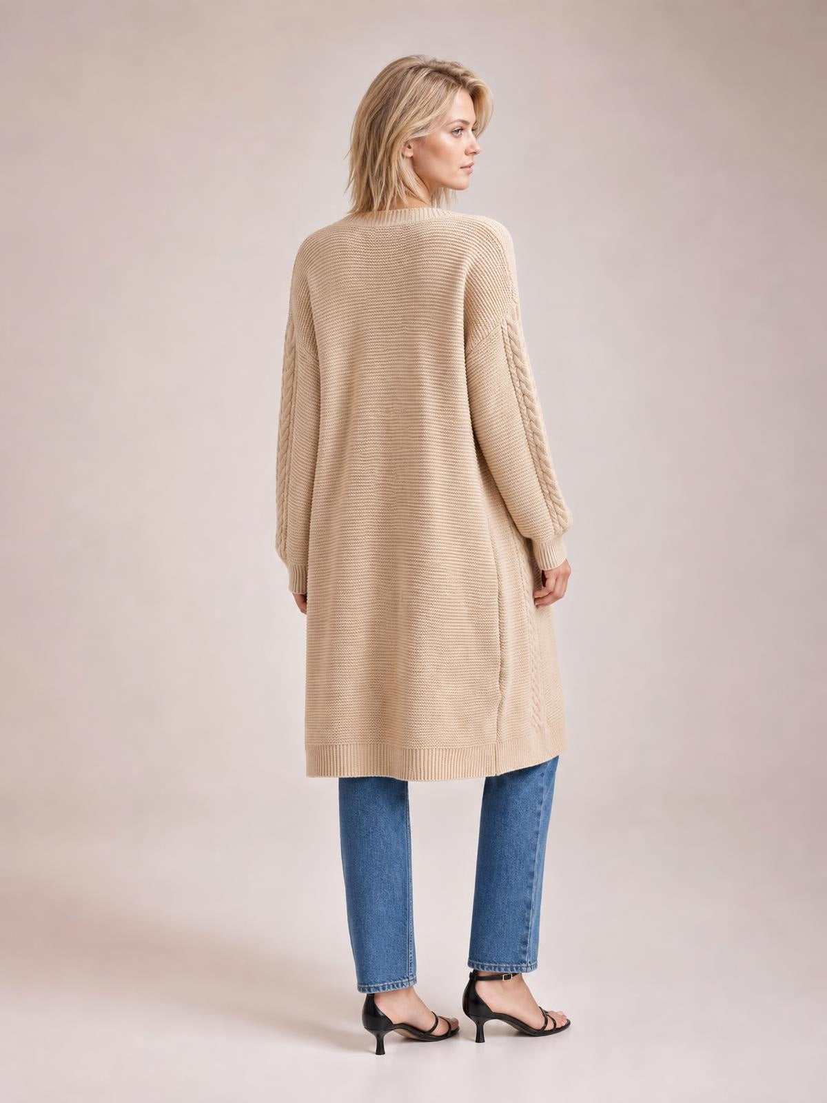 Model shown from the back wears a beige cable-knit open-front longline cardigan with a relaxed silhouette, ribbed hem and cuffs, and braided sleeves.