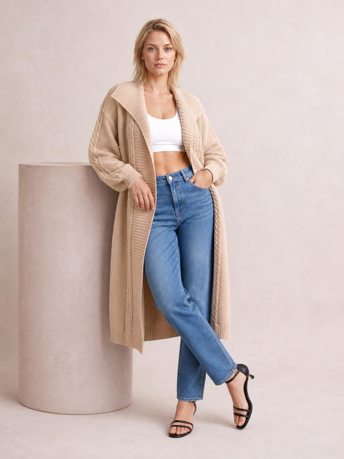 A model wears a beige cable-knit open-front longline cardigan with ribbed cuffs and hem, relaxed sleeves, and a flowing silhouette while leaning casually.