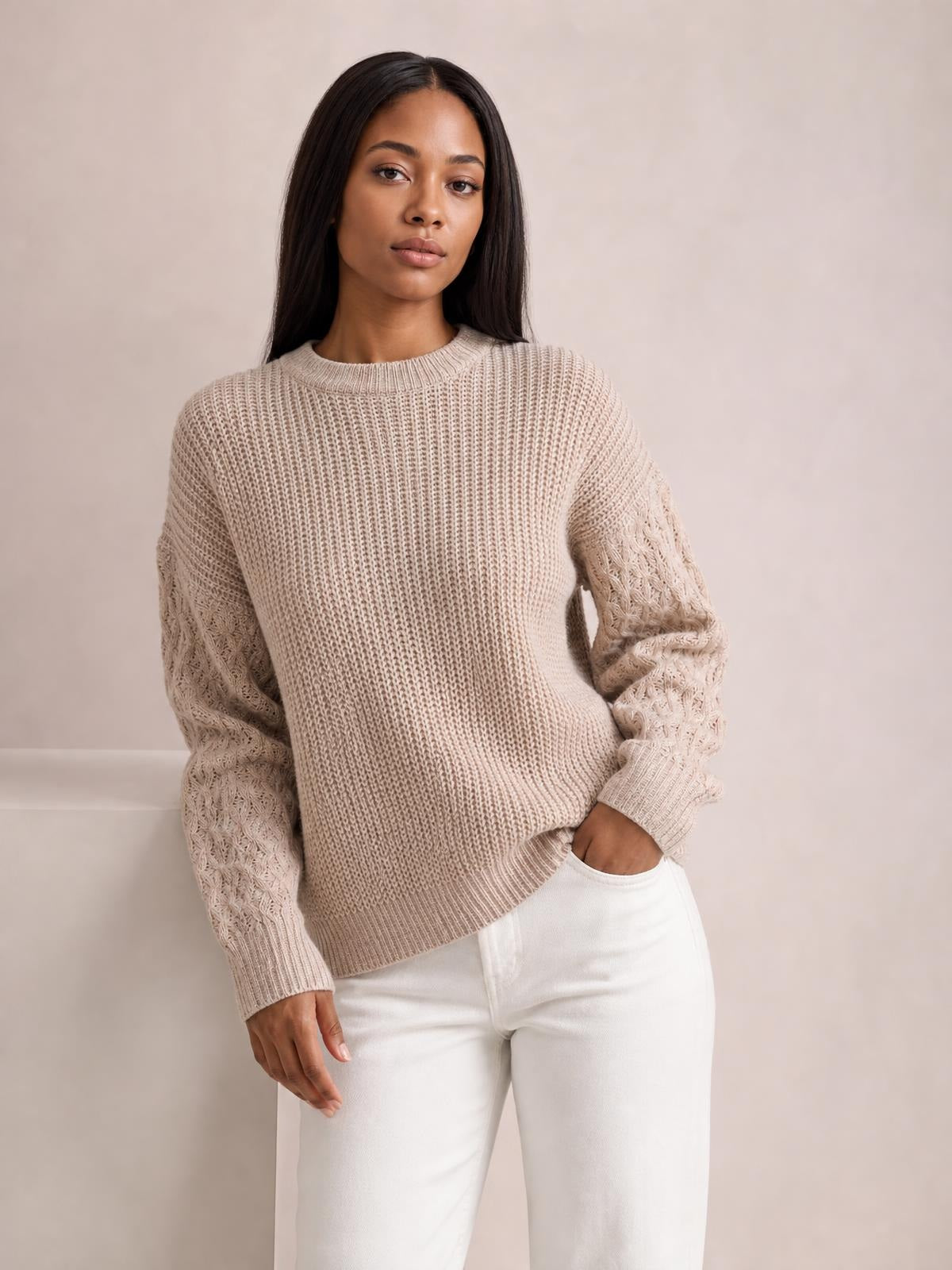 Model poses in a beige cable-knit round-neck sweater with dropped shoulders, textured sleeves, ribbed cuffs and hem, and a relaxed silhouette.