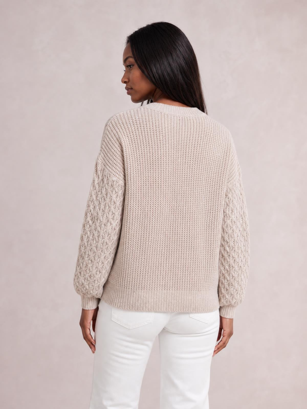 A model looks over her shoulder wearing a cream cable-knit round-neck sweater with dropped shoulders, relaxed fit, textured sleeves, and ribbed cuffs and hem.
