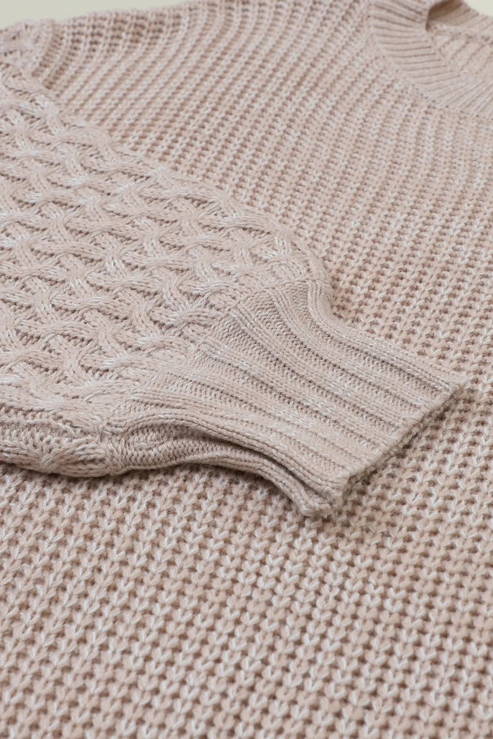 Close-up of a beige cable-knit sweater showing its textured knit construction and ribbed sleeve cuff in a soft, relaxed silhouette.