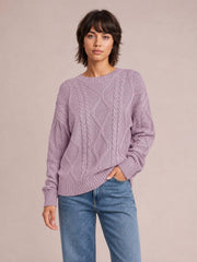 Model wearing a lavender cable-knit round neck sweater with long sleeves, ribbed cuffs and hem, relaxed silhouette, and textured diamond and braided construction.