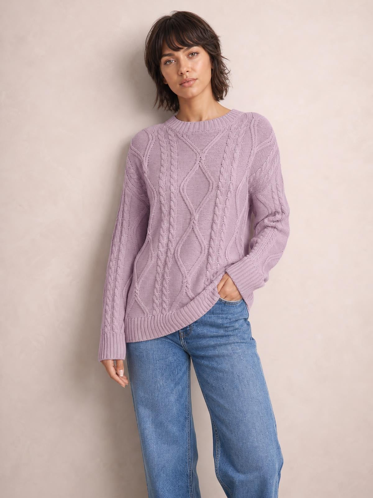 Model wearing a lilac cable-knit round-neck sweater with long sleeves, ribbed trim, and a relaxed silhouette, posing with one hand in her jeans pocket.