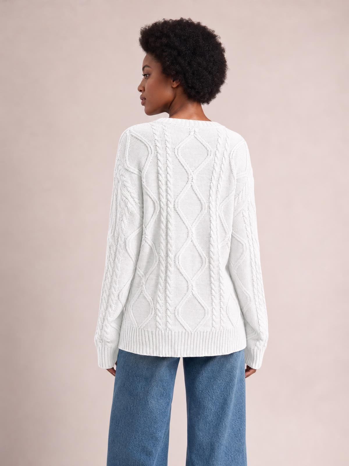 Model stands turned away in a white cable-knit round neck sweater with long sleeves, ribbed cuffs and hem, and a relaxed silhouette.