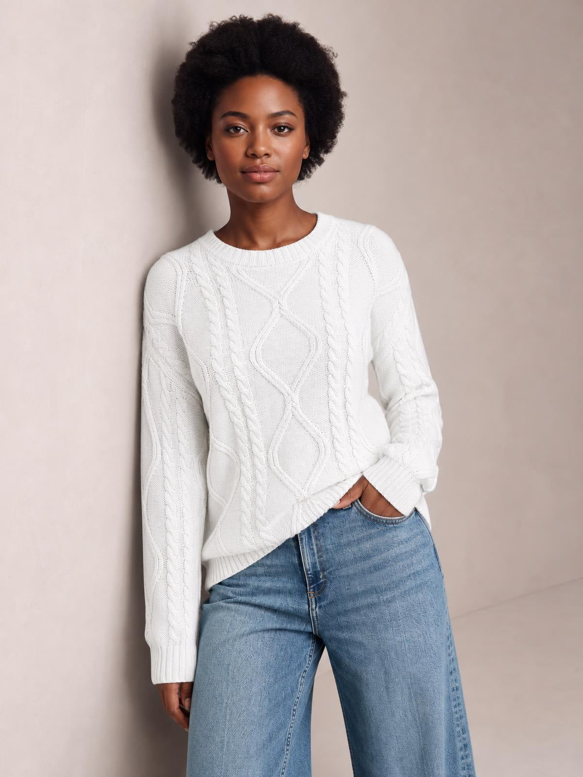 Model poses in a white cable-knit round neck sweater with long sleeves, ribbed cuffs and hem, and a relaxed silhouette.