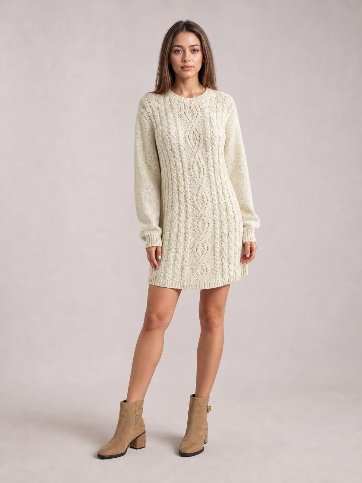 Model stands in a cream cable knit sweater mini dress with a straight silhouette, long sleeves, crew neckline, and detailed braided knit texture.