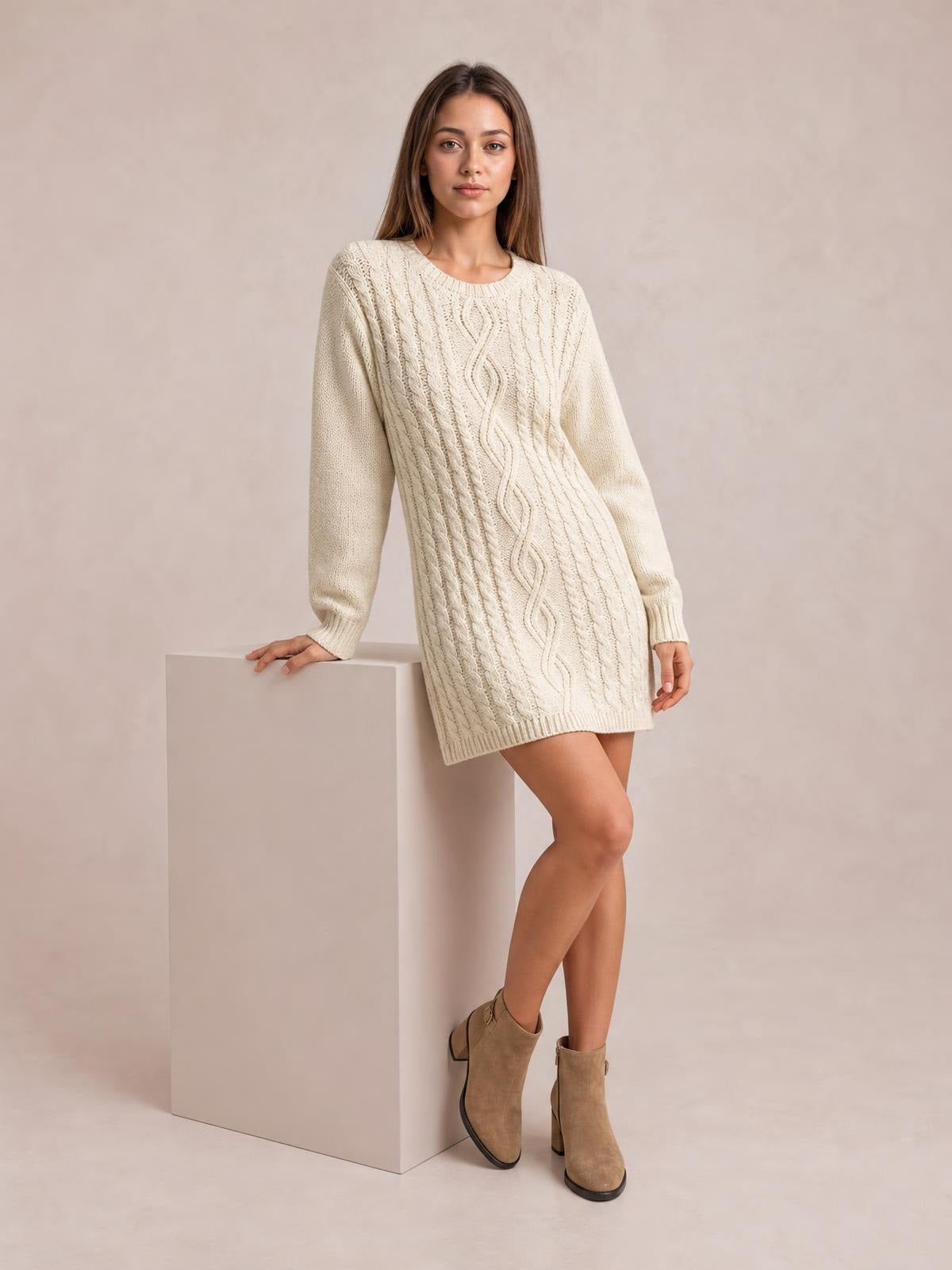 Model poses in a cream cable-knit sweater mini dress with long sleeves, ribbed trim, visible seam detail, and a straight silhouette.