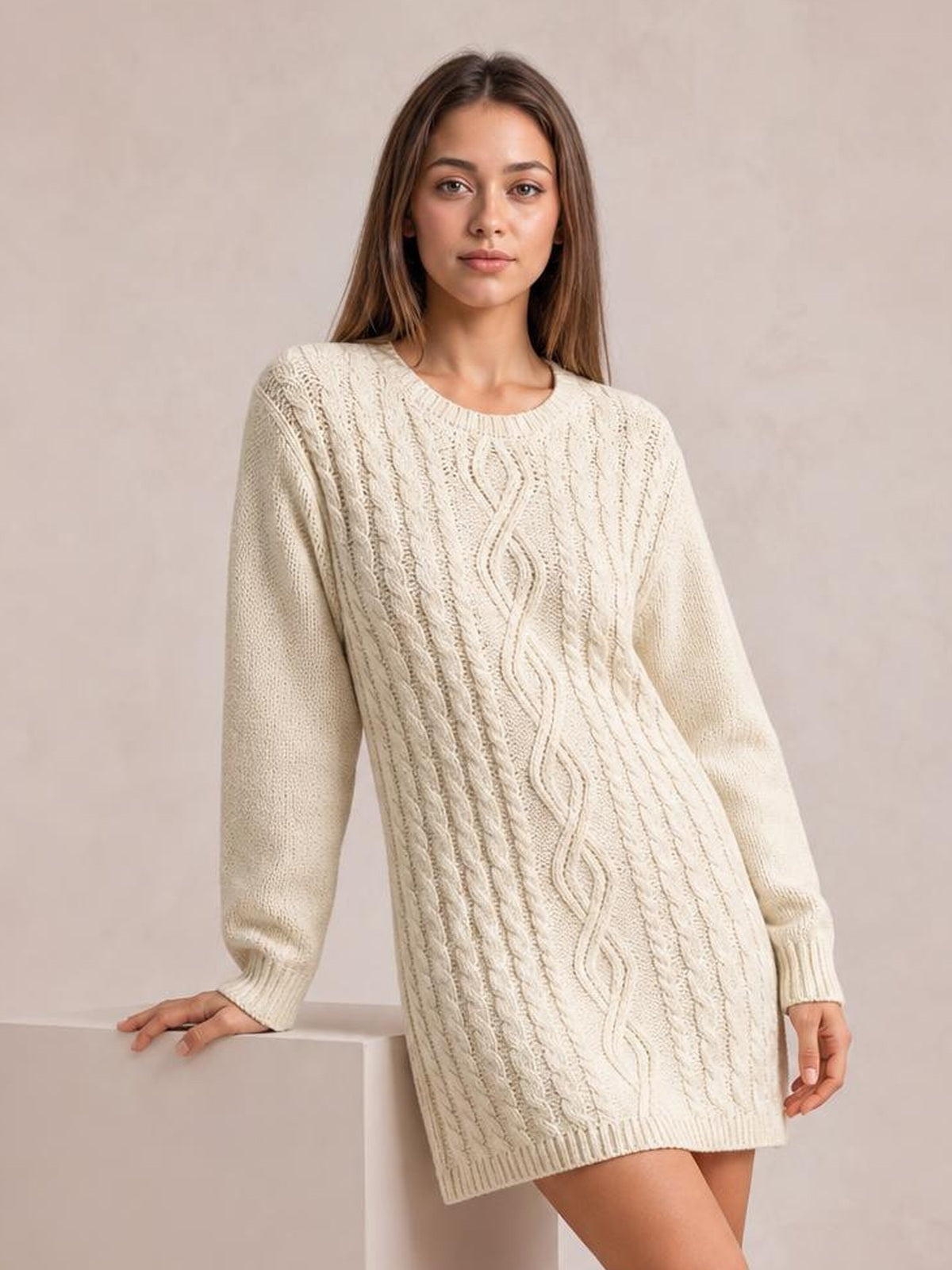 Model poses in a cream cable-knit sweater mini dress with a relaxed straight silhouette, round neckline, long sleeves, ribbed cuffs, and hem.