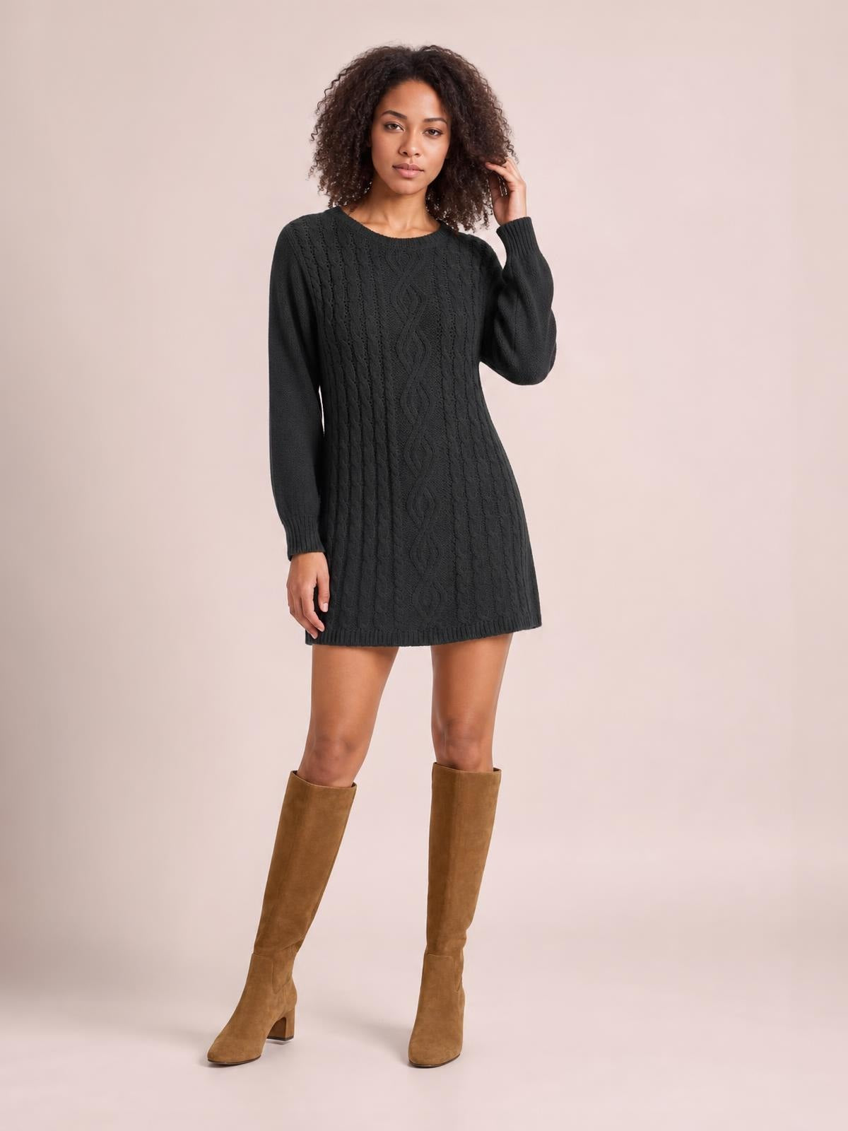 Model poses in a black cable-knit sweater mini dress with long sleeves, a fitted silhouette, round neckline, braided knit detail, and ribbed hem.