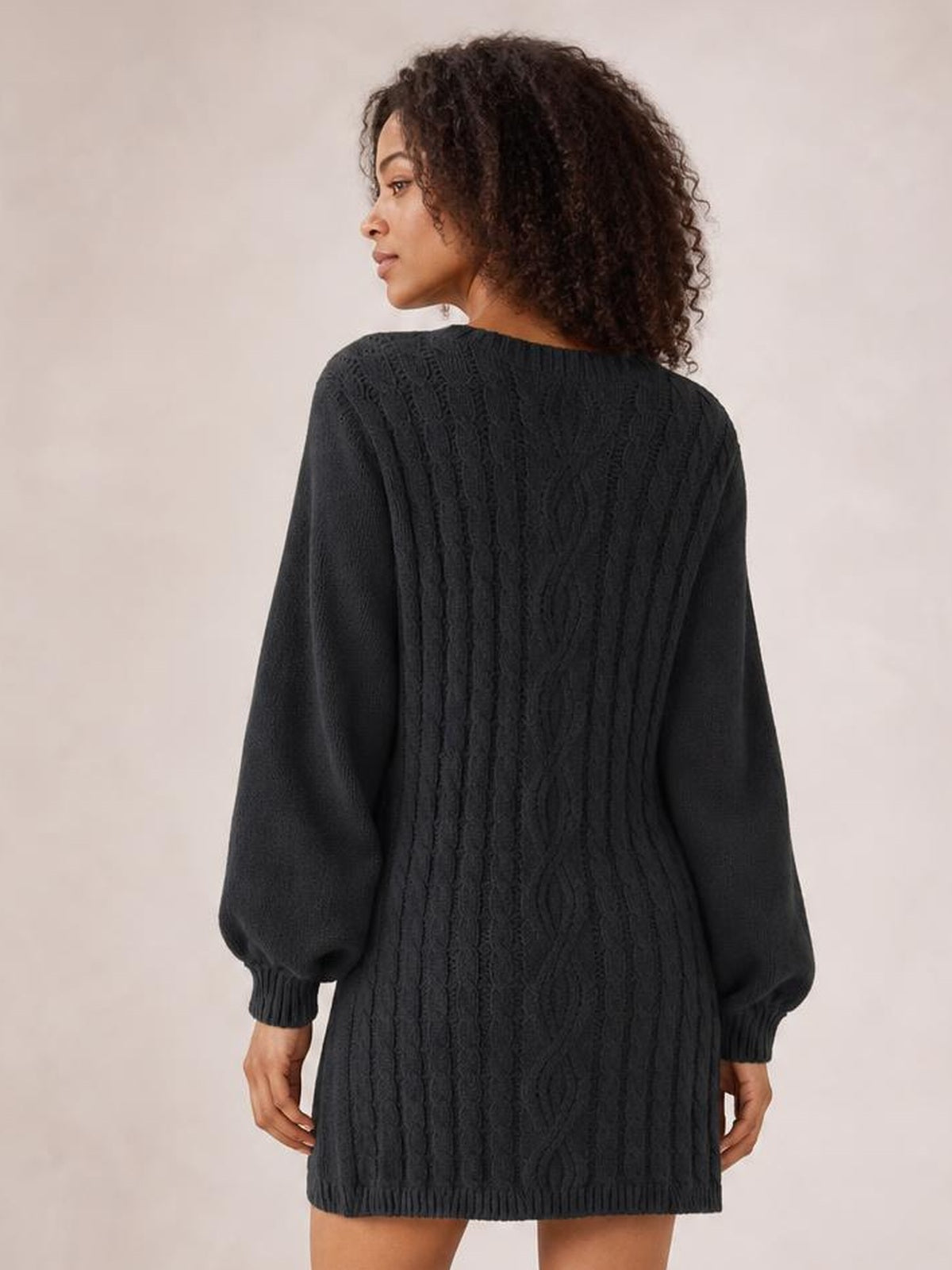 Model looks over her shoulder while wearing a black cable-knit sweater mini dress with long sleeves, ribbed cuffs and hem, and a relaxed straight silhouette.