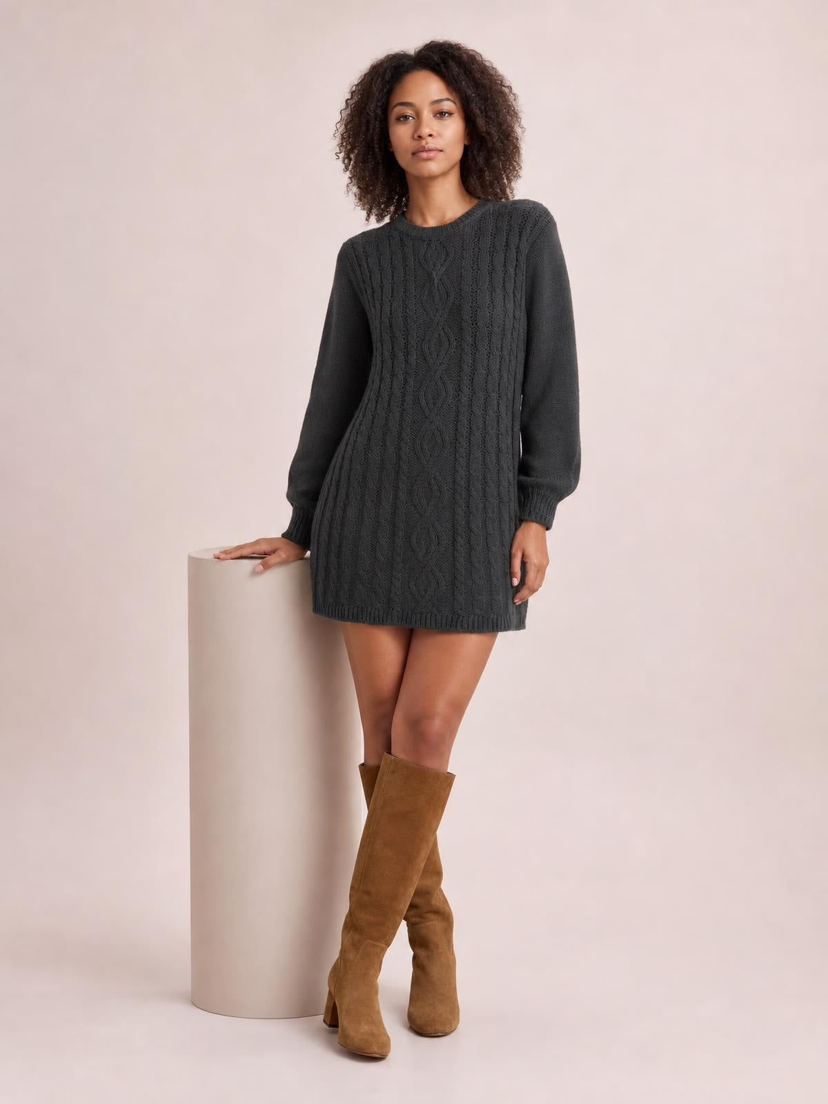 A model poses in a charcoal cable knit sweater mini dress with long sleeves, a round neckline, straight silhouette, and ribbed cuffs and hem.