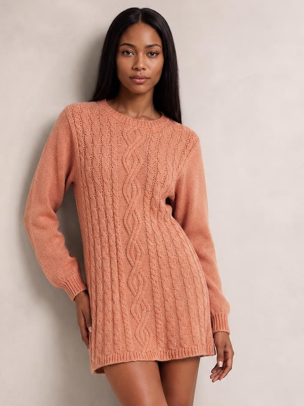 A model stands wearing a coral cable-knit sweater mini dress with long sleeves, crew neckline, braided cable panels, ribbed cuffs and hem, and a relaxed straight silhouette.