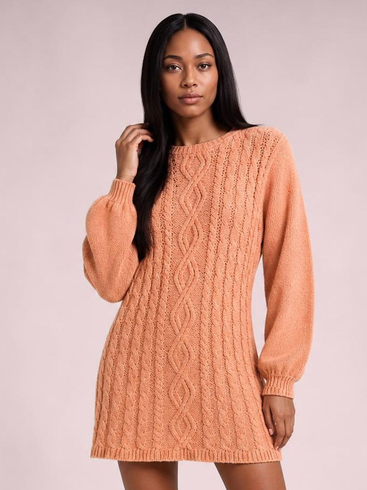 Model poses in a peach cable-knit sweater mini dress with long sleeves, ribbed crew neckline and cuffs, diamond-patterned front, and straight silhouette.