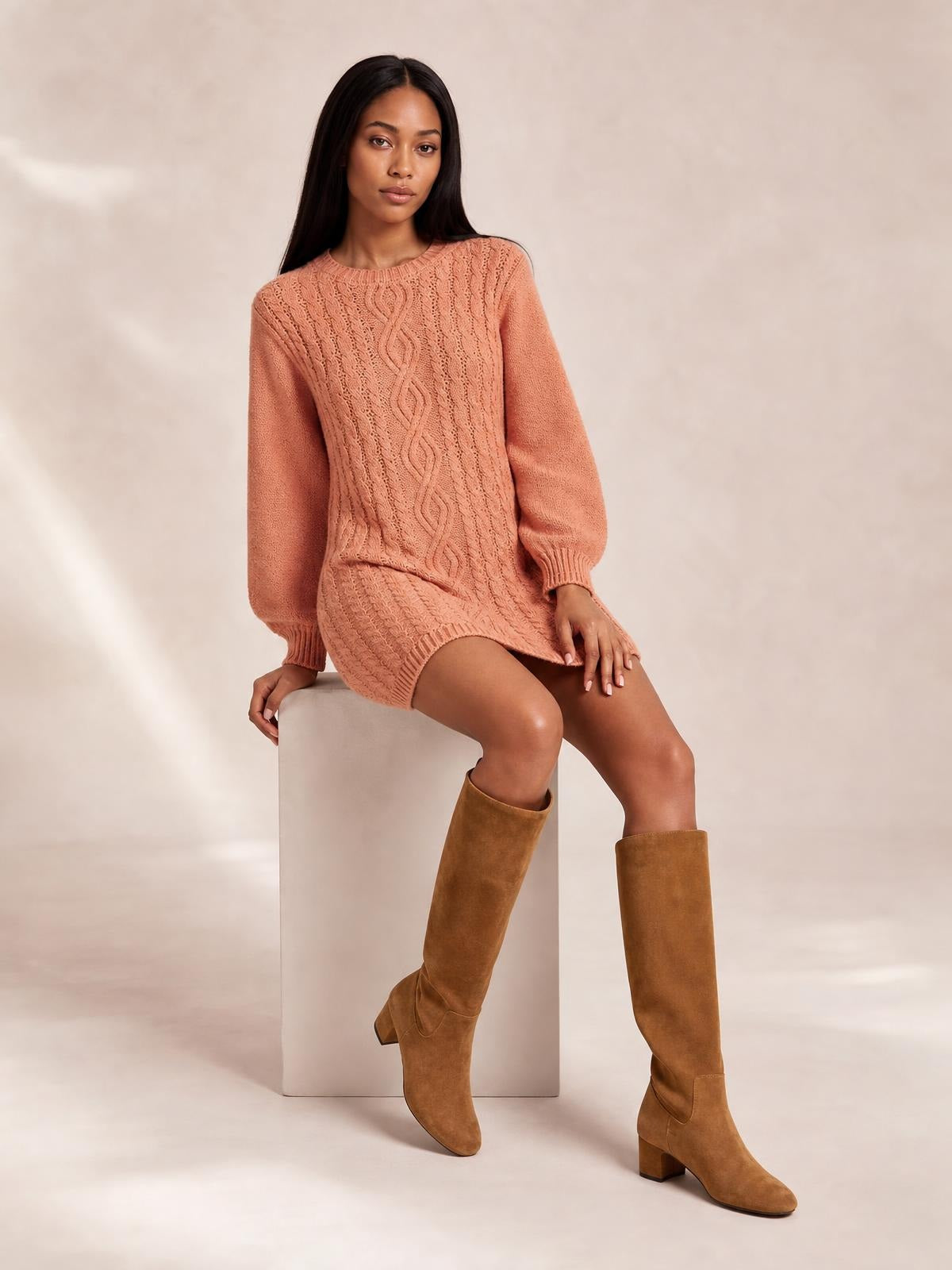 A seated model wears a peach cable-knit sweater mini dress with long sleeves, ribbed cuffs and hem, a crew neckline, and a relaxed straight silhouette.