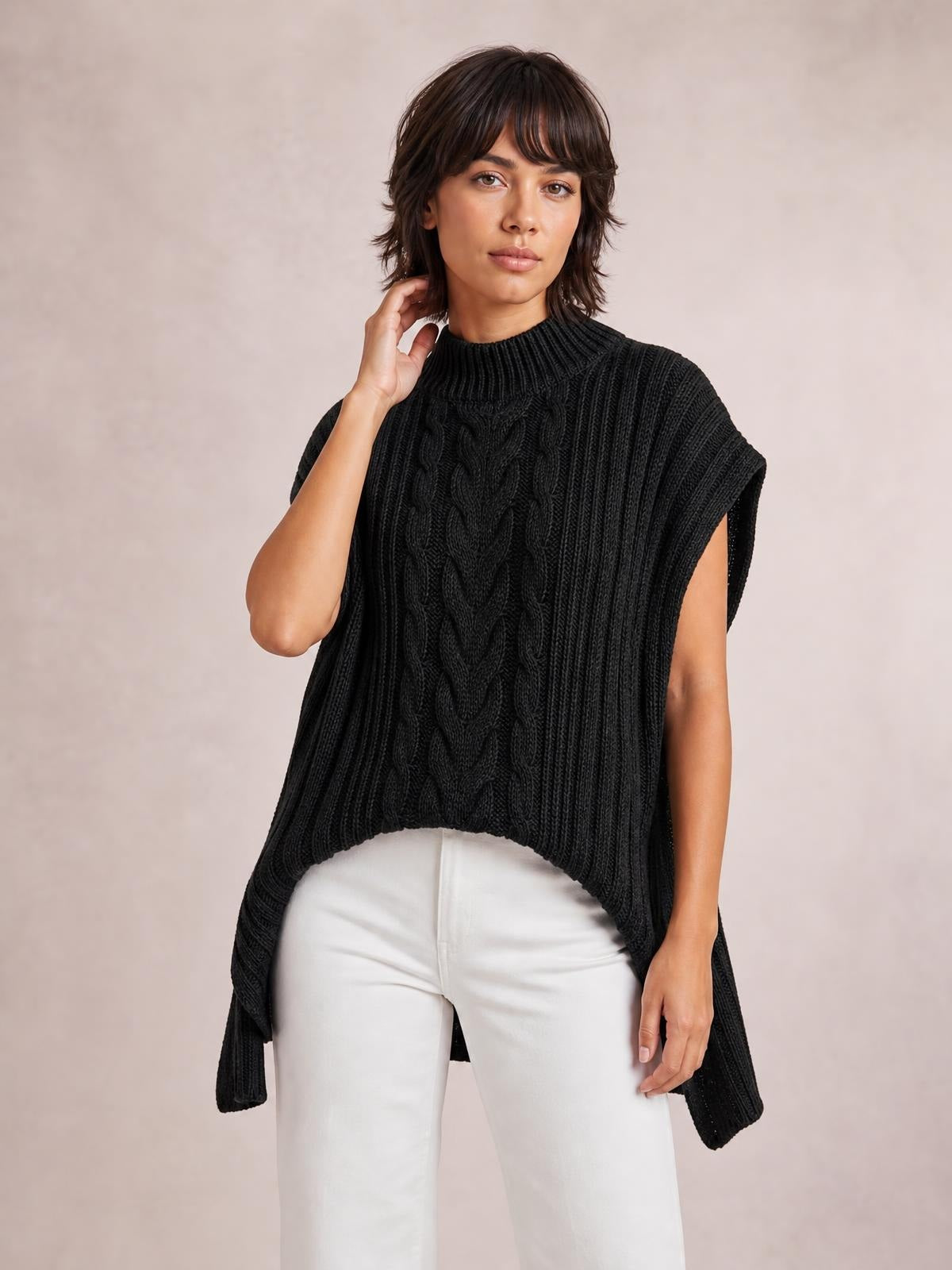 A model wears a black cable-knit sweater vest with a ribbed high neckline, sleeveless oversized silhouette, and elongated side panels, posing with one hand near her neck.
