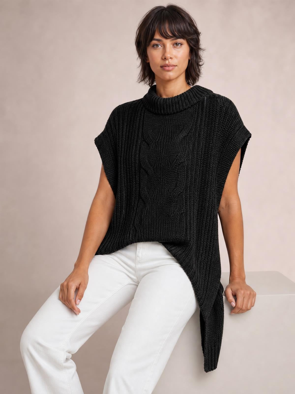 Model seated in a black cable-knit sweater vest with a ribbed high neckline, sleeveless dropped-shoulder silhouette, and asymmetrical long side hem.