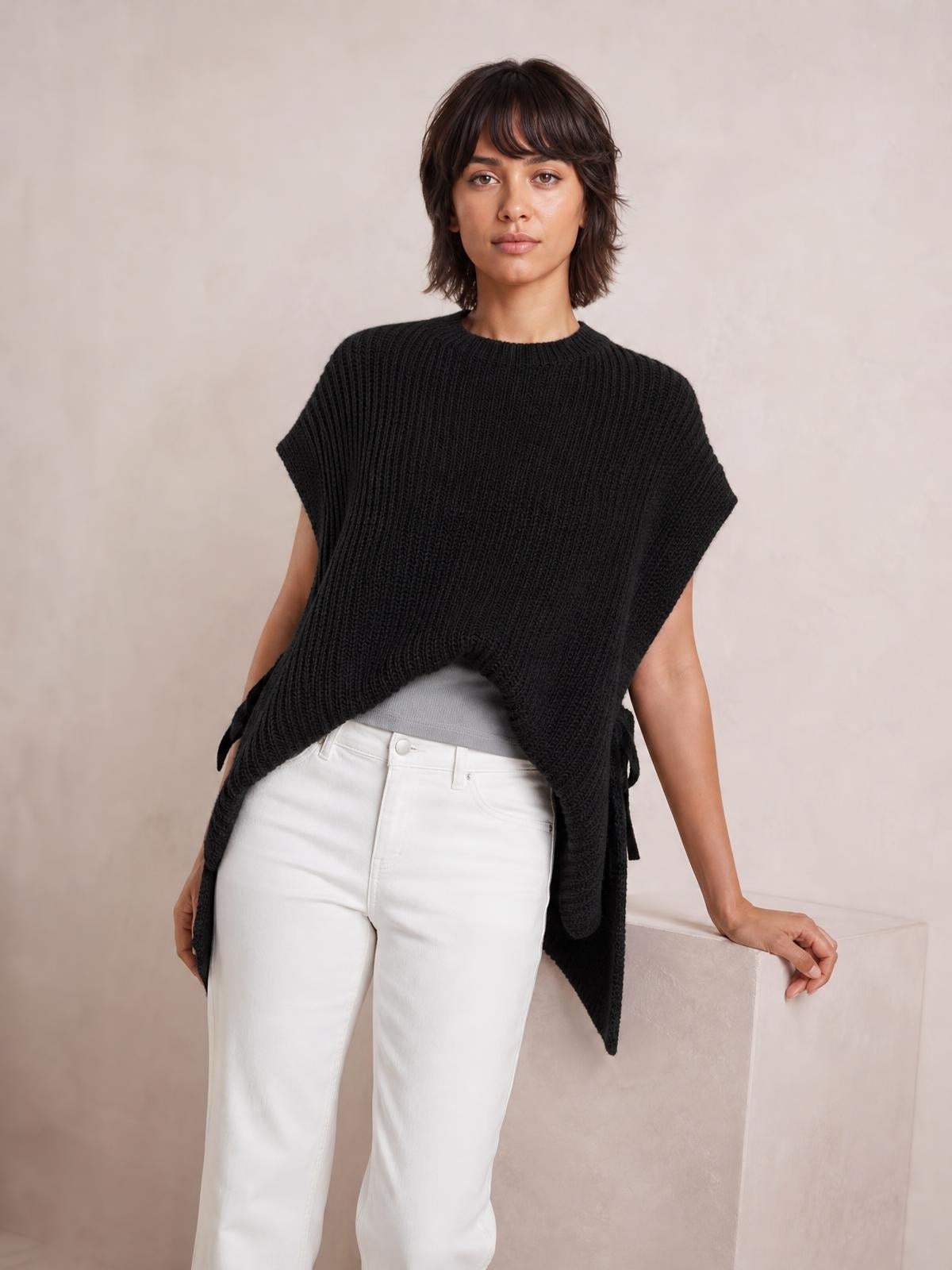 A model stands in a black cable-knit sweater vest with a round neck, wide armholes, oversized silhouette, and pointed side hems over a gray top and white pants.