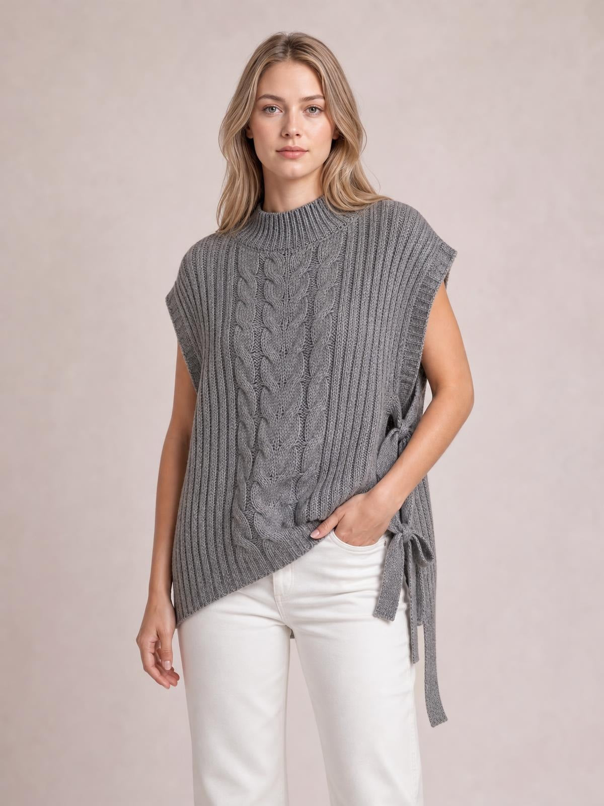 Model wearing a gray cable knit sweater vest with a high ribbed neckline, sleeveless cut, relaxed silhouette, and side tie detail over white pants.