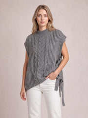Model wearing a gray cable knit sweater vest with a high ribbed neckline, sleeveless cut, relaxed silhouette, and side tie detail over white pants.