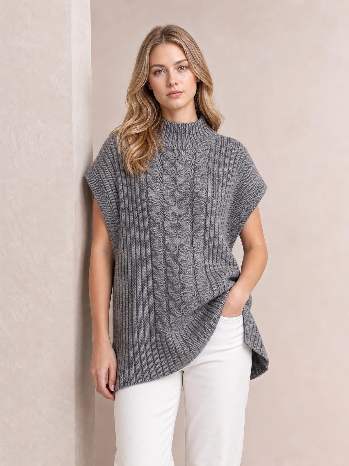 Model wearing a gray cable-knit sweater vest with a high neck, sleeveless cut, relaxed silhouette, textured ribbed sides, and a longer curved hem.