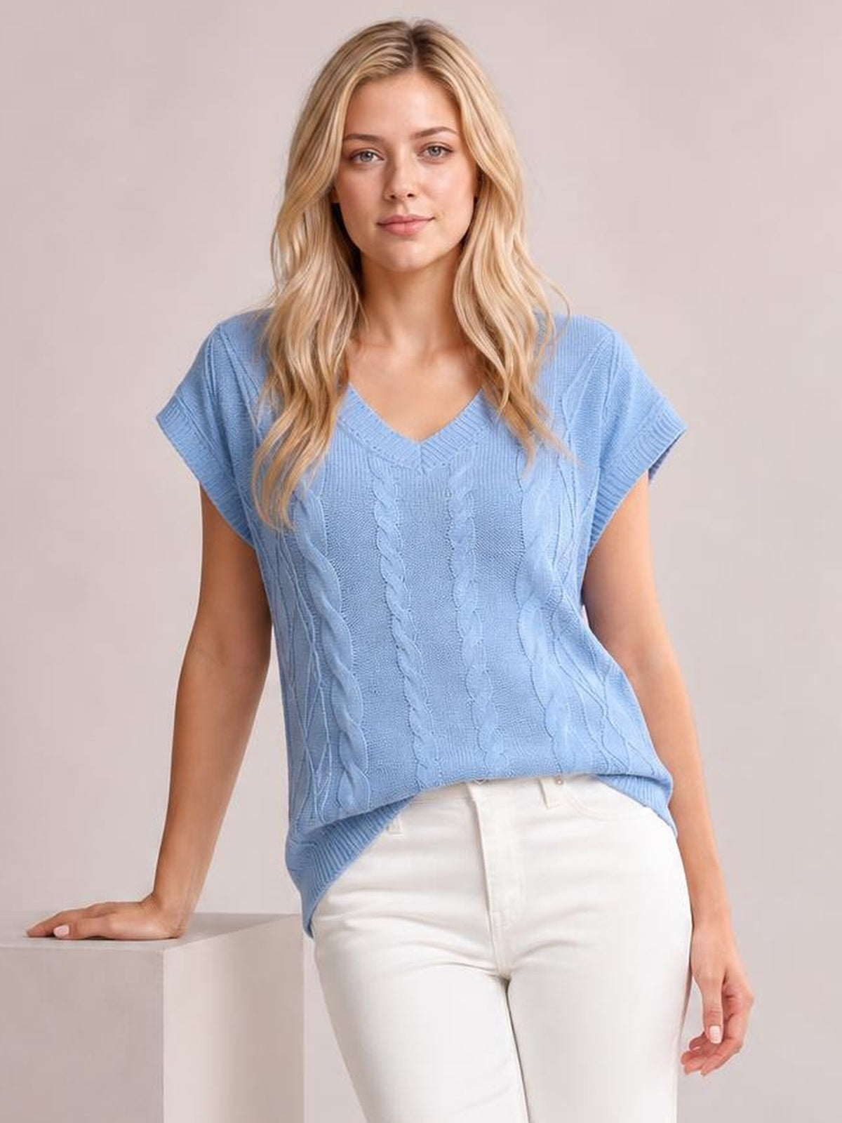 Model poses in a light blue cable-knit V-neck short sleeve top with a relaxed silhouette, ribbed trim, and a slightly curved hem over white pants.