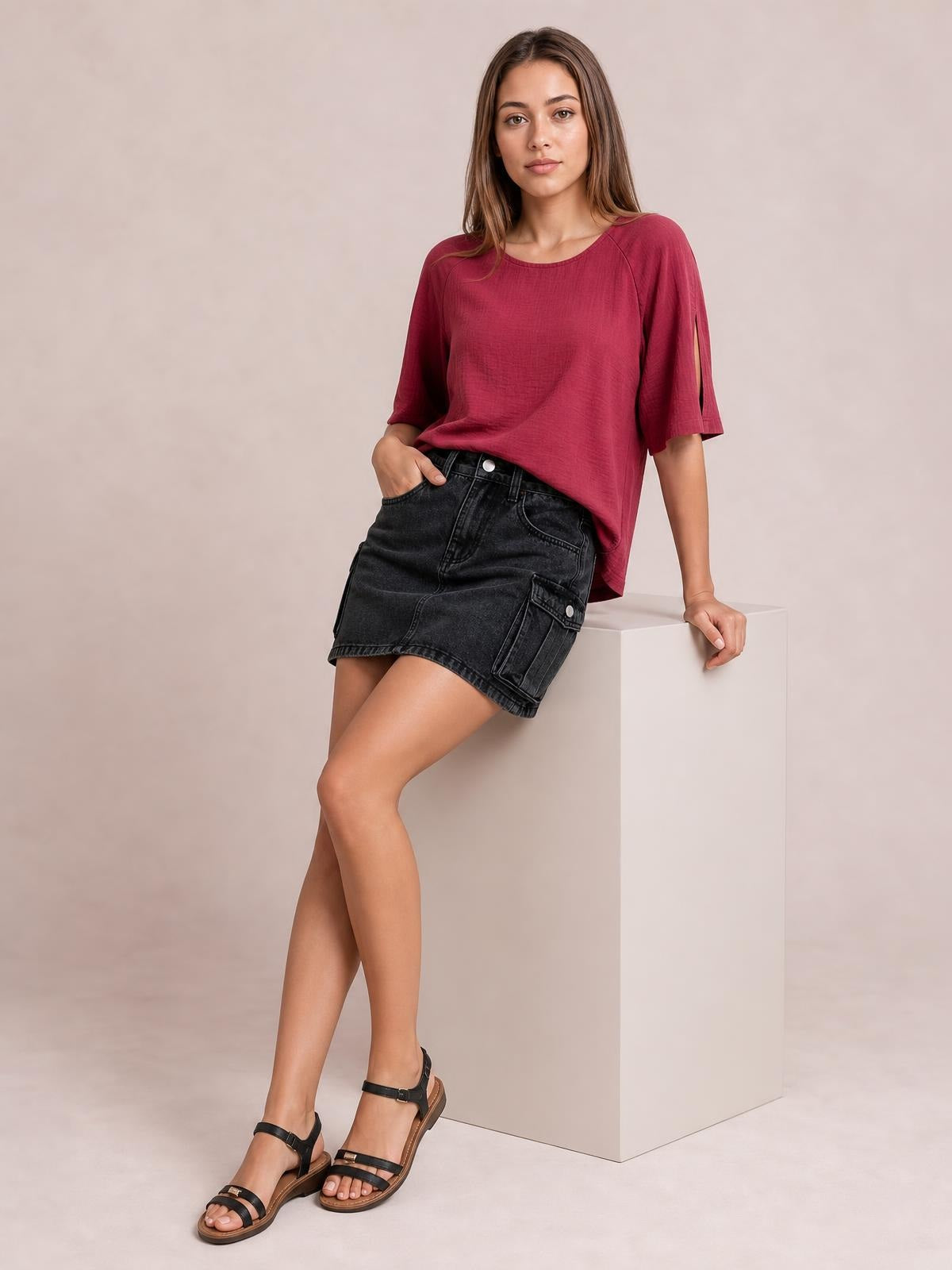 Model leans against a cube in a black denim cargo skirt with a high-rise waist, button closure, front pockets, side utility pocket, and straight mini silhouette.