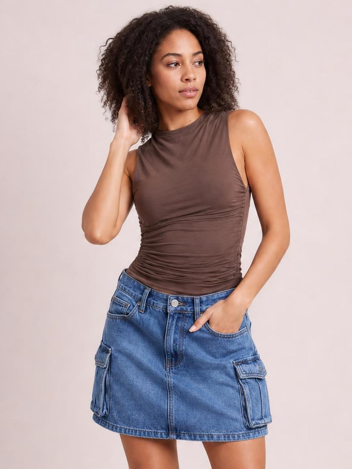 Model poses with one hand in a blue denim cargo skirt’s pocket, showing a high waist, straight mini silhouette, front fly, belt loops, and flap cargo pockets.