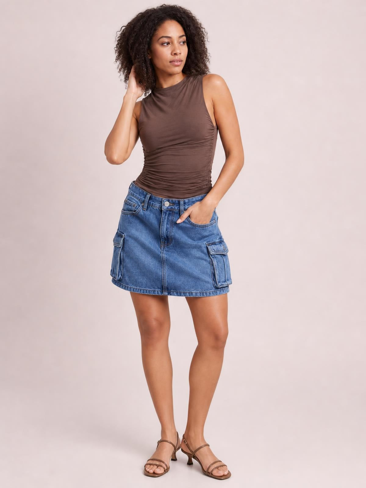 Model in a relaxed standing pose wears a blue denim cargo skirt with a straight mini silhouette, belt loops, front pockets, and large flap cargo pockets.
