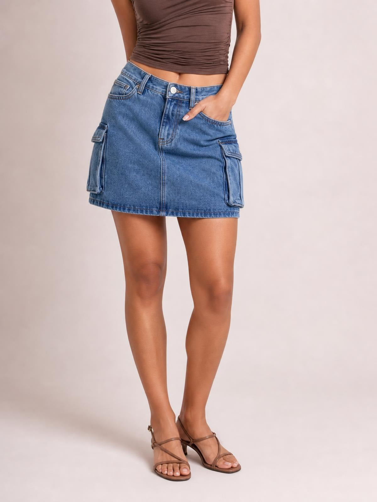 Model wears a blue denim cargo skirt with a straight mini silhouette, belt loops, zip fly, front pockets, and large flap cargo pockets while standing with one hand tucked in.