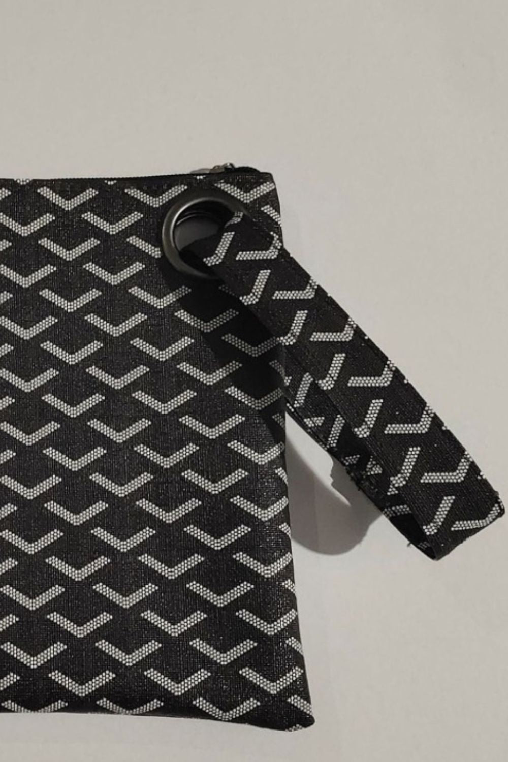 A black PU leather wristlet features a rectangular silhouette, repeating white chevron pattern, top zipper, and attached wrist strap with a metal ring.