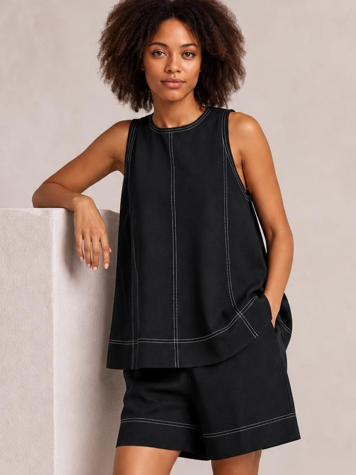 A model poses in a relaxed black denim tank top and shorts set with an oversized silhouette, contrast stitching, seam details, and a pocketed design.