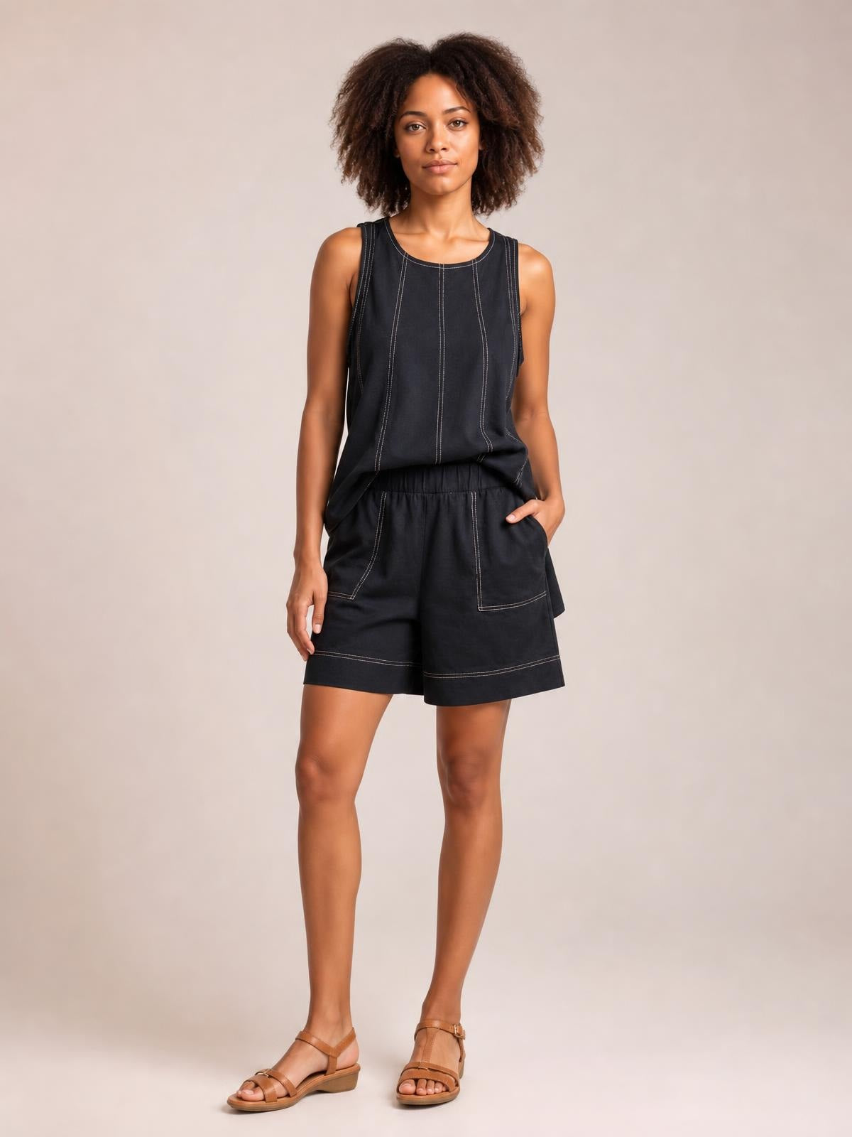 Model wearing a relaxed black denim tank top and shorts set with an oversized silhouette, contrast stitching, seam details, and pockets, posing with one hand tucked in.