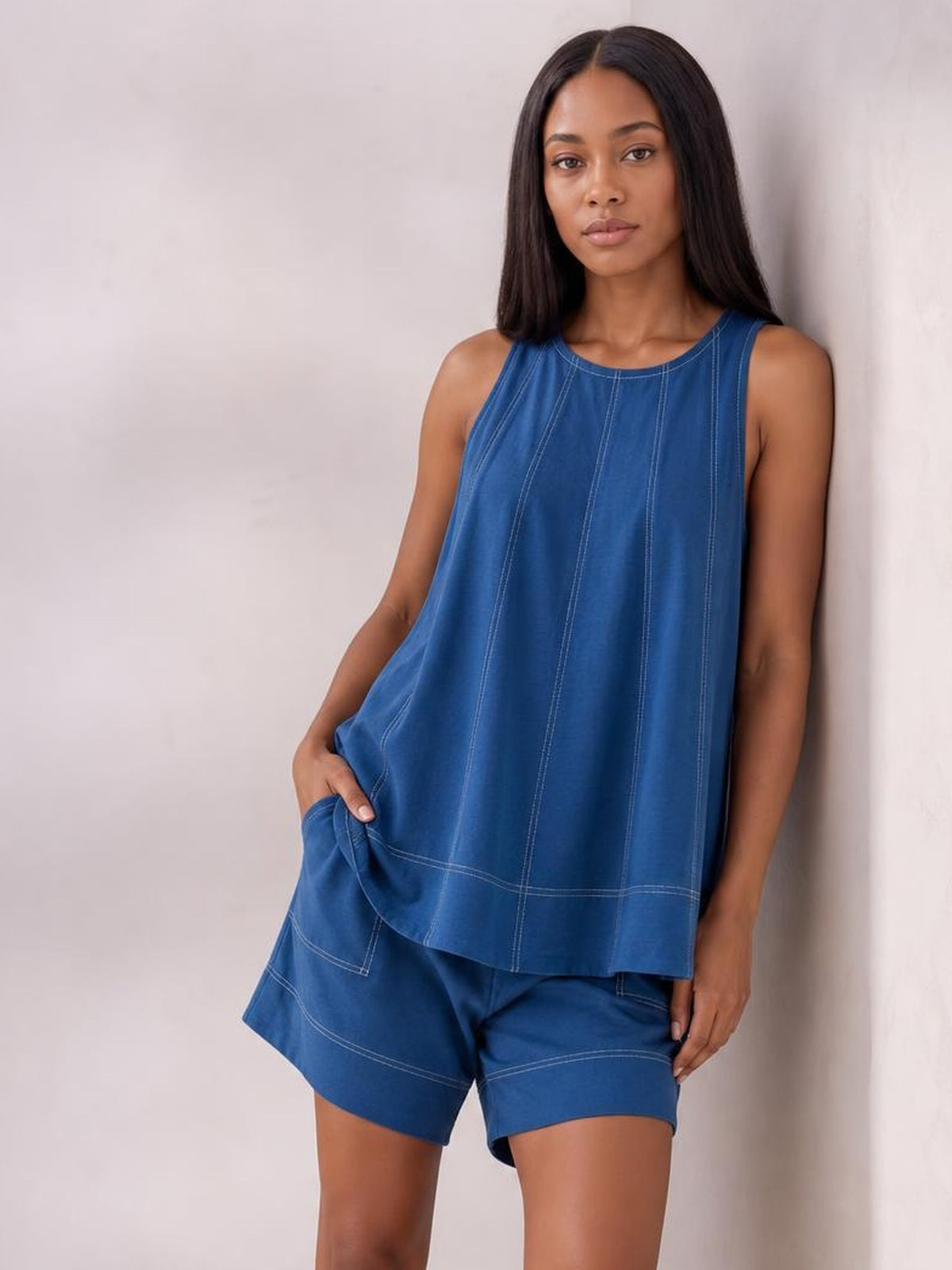 Model poses with one hand in the pocket, wearing a relaxed blue denim tank top and shorts set with an oversized silhouette and contrast stitching.