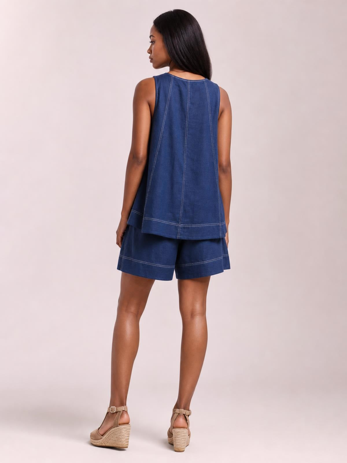 A model stands in a rear three-quarter pose wearing a relaxed blue denim tank top and loose shorts set with oversized silhouettes and contrasting seam stitching.