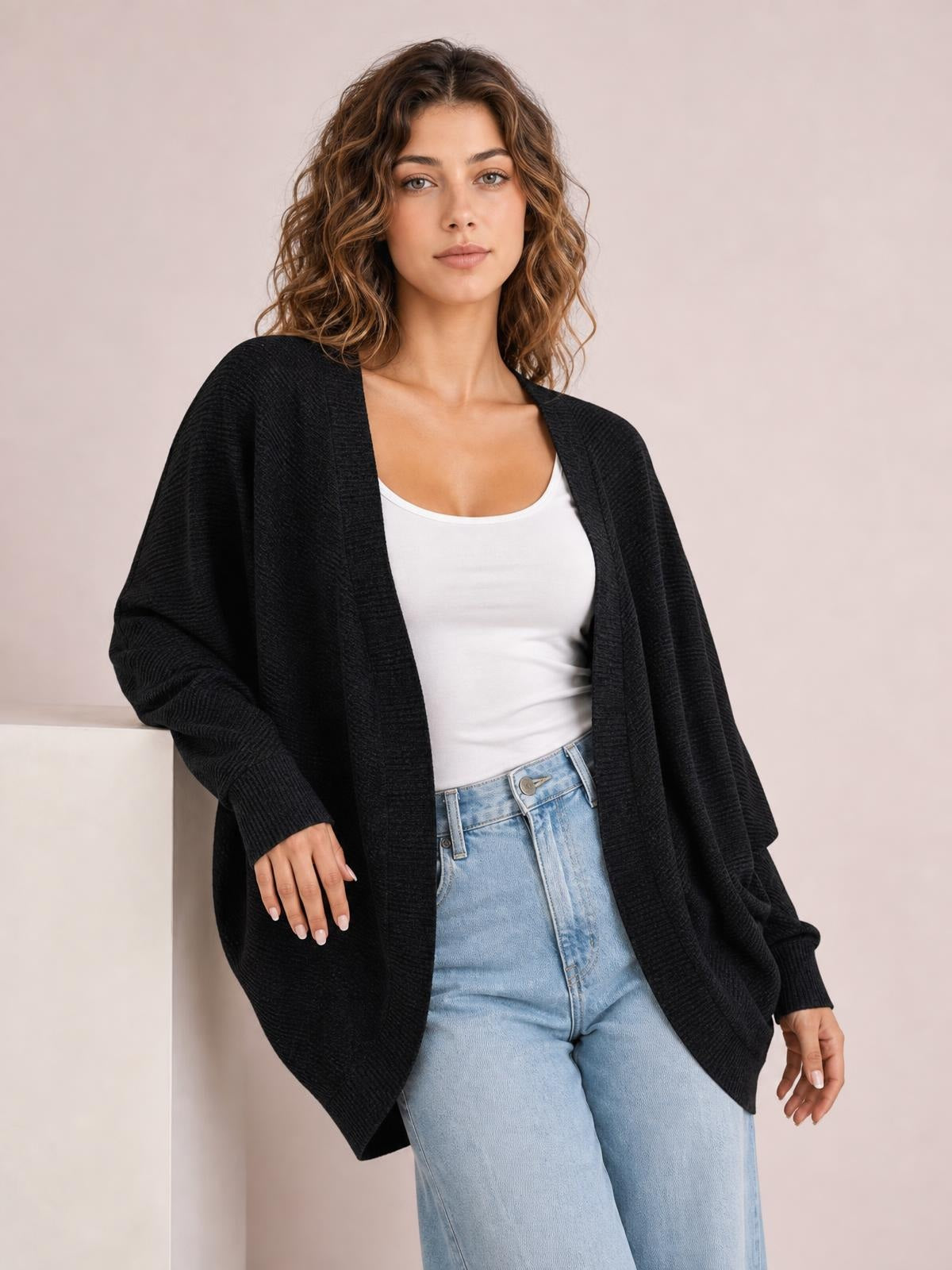 A woman poses in a black knit open front cardigan with a relaxed draped silhouette, long sleeves, and ribbed cuffs over a white tank top.