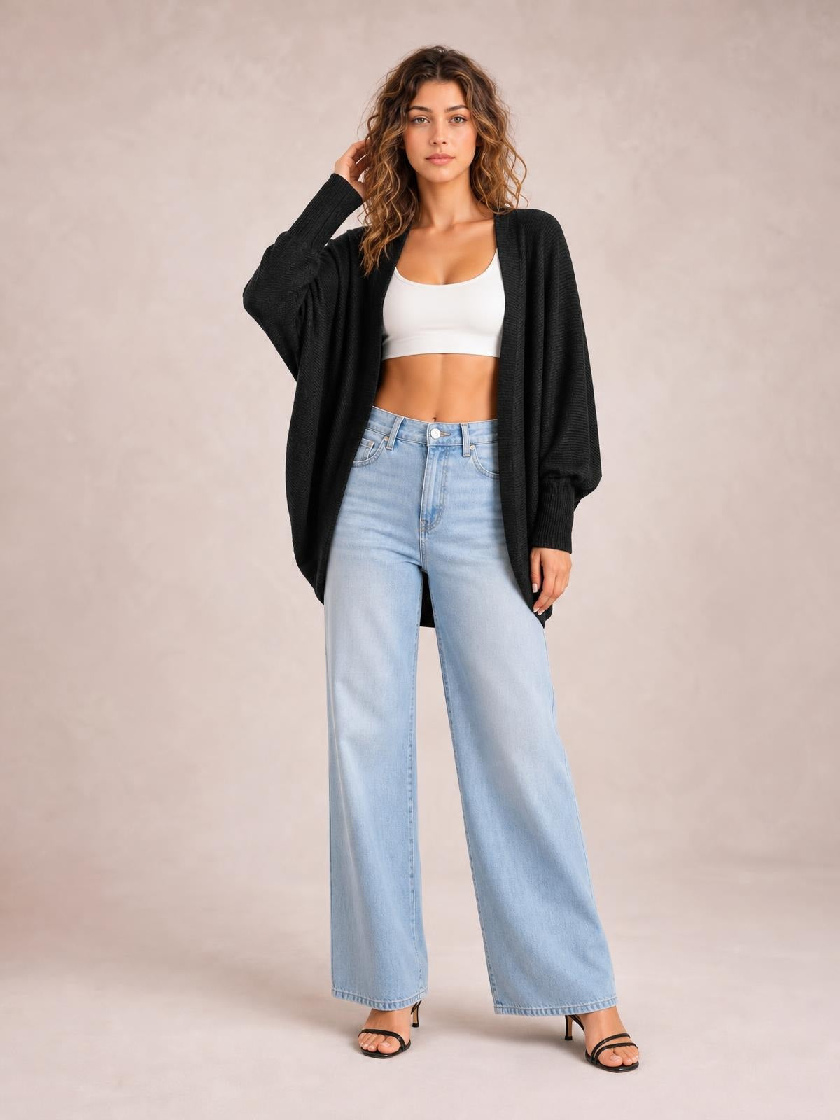 Model poses in a black knit open-front cardigan with long sleeves, ribbed cuffs, and a relaxed draped silhouette over a white crop top.